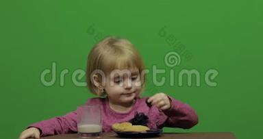 女孩坐在桌子旁吃巧克力、饼干和饮料可可。 快乐的三岁女孩。 可爱的女孩微笑